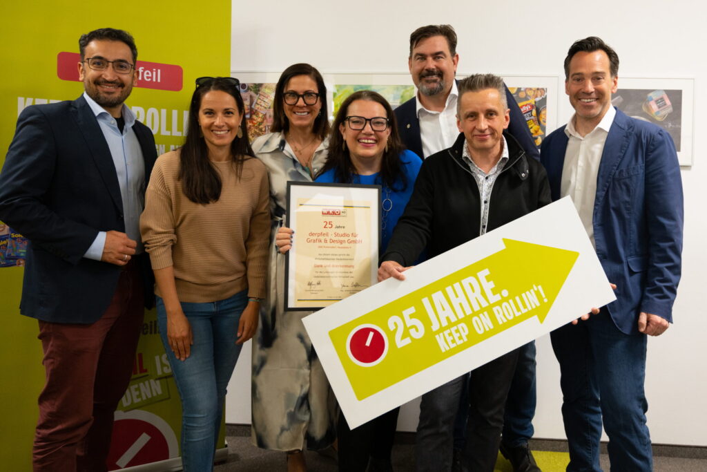 Sechs Personen stehen lächelnd nebeneinander in einem hellen Raum. Eine Frau in der Mitte hält eine gerahmte Ehrenurkunde der Wirtschaftskammer Niederösterreich zum 25-jährigen Bestehen des Studios „derpfeil“. Ein Mann davor präsentiert ein Schild mit der Aufschrift „25 JAHRE. KEEP ON ROLLIN’!“.