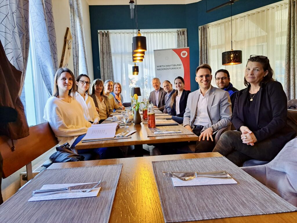 Eine Gruppe von elf Personen sitzt an einer langen Tafel in einem hellen Restaurant mit blauen Akzentwänden. Im Hintergrund ist ein Roll-up-Banner des Wirtschaftsbundes Niederösterreich zu sehen. Die Teilnehmer blicken lächelnd in die Kamera, während auf dem Tisch Getränke und Gedecke vorbereitet sind.