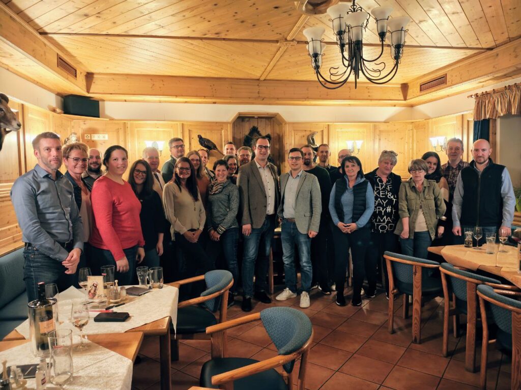 Ein Gruppenfoto von etwa 25 Personen bei einem Unternehmerstammtisch in einem rustikal eingerichteten Gasthaus mit Holzvertäfelung. Die Teilnehmer stehen verteilt im Raum und lächeln in die Kamera; im Vordergrund sind gedeckte Tische mit Gläsern erkennbar.