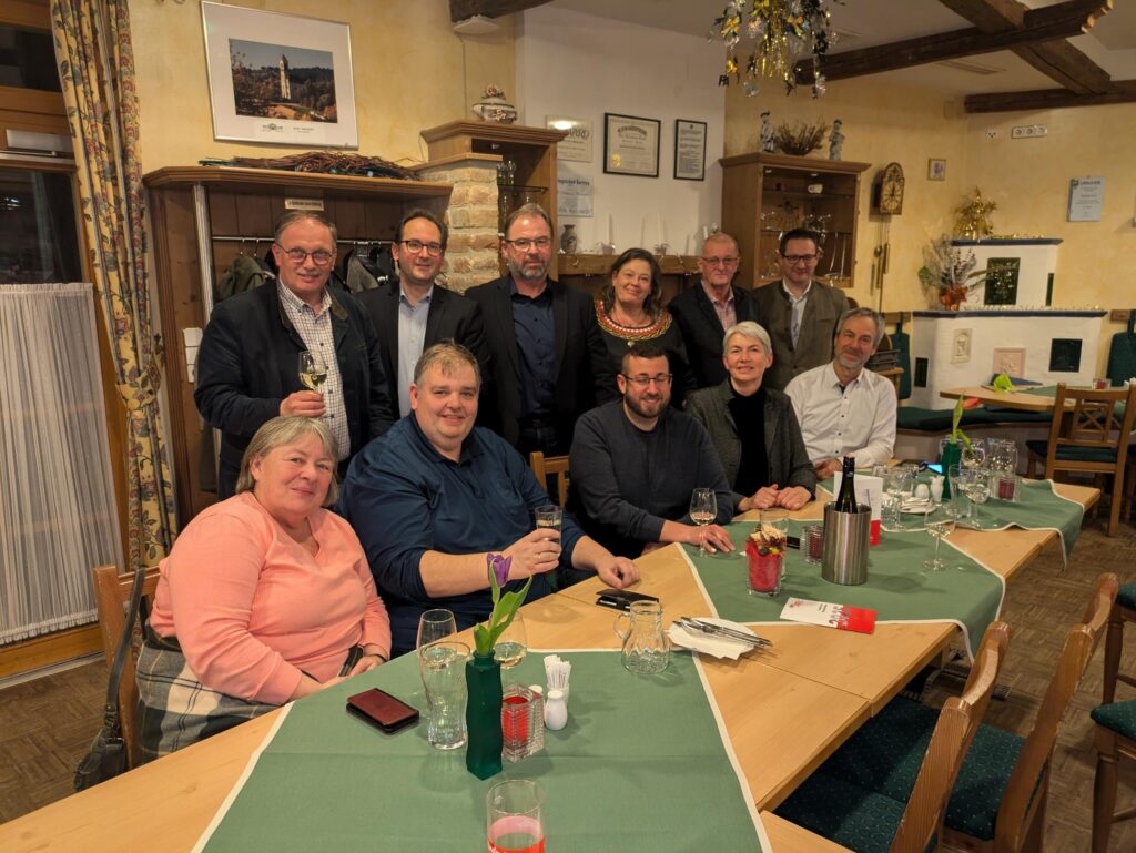Eine Gruppe von elf Personen beim Stammtisch des Hollabrunner Wirtschaftsbundes. Fünf Personen sitzen an einem mit grünen Läufern gedeckten langen Tisch, sechs weitere stehen dahinter in einem gemütlich eingerichteten Gasthaus mit hellgelben Wänden und Holzdeckenbalken.
