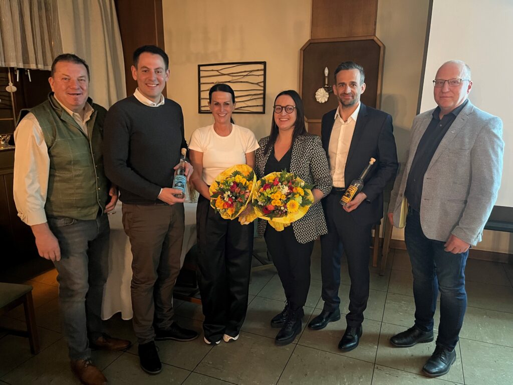 Gruppenfoto von sechs Personen beim Wirtschaftstreff in Hollenthon. Zwei Frauen in der Mitte halten gelbe Blumensträuße, flankiert von zwei Männern, die jeweils eine gläserne Flasche präsentieren. Die Gruppe lächelt in einem gemütlichen Gastraum in die Kamera.