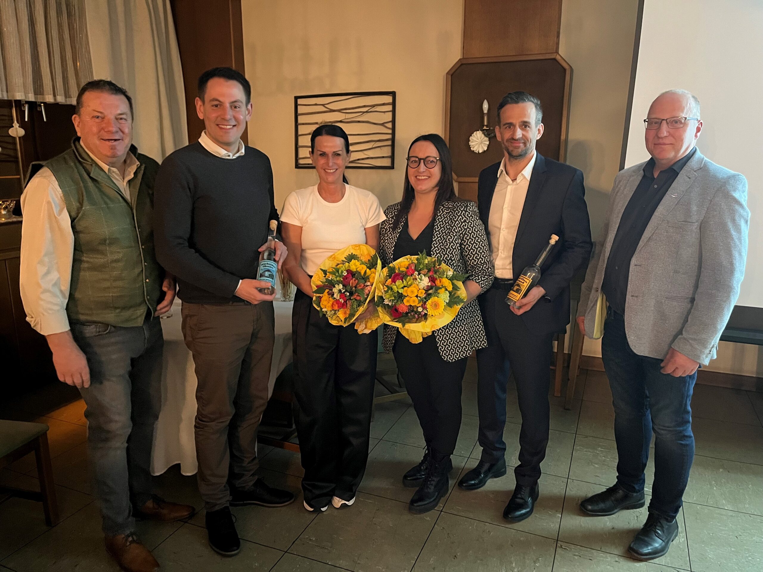 Gruppenfoto von sechs Personen beim Wirtschaftstreff in Hollenthon. Zwei Frauen in der Mitte halten gelbe Blumensträuße, flankiert von zwei Männern, die jeweils eine gläserne Flasche präsentieren. Die Gruppe lächelt in einem gemütlichen Gastraum in die Kamera.