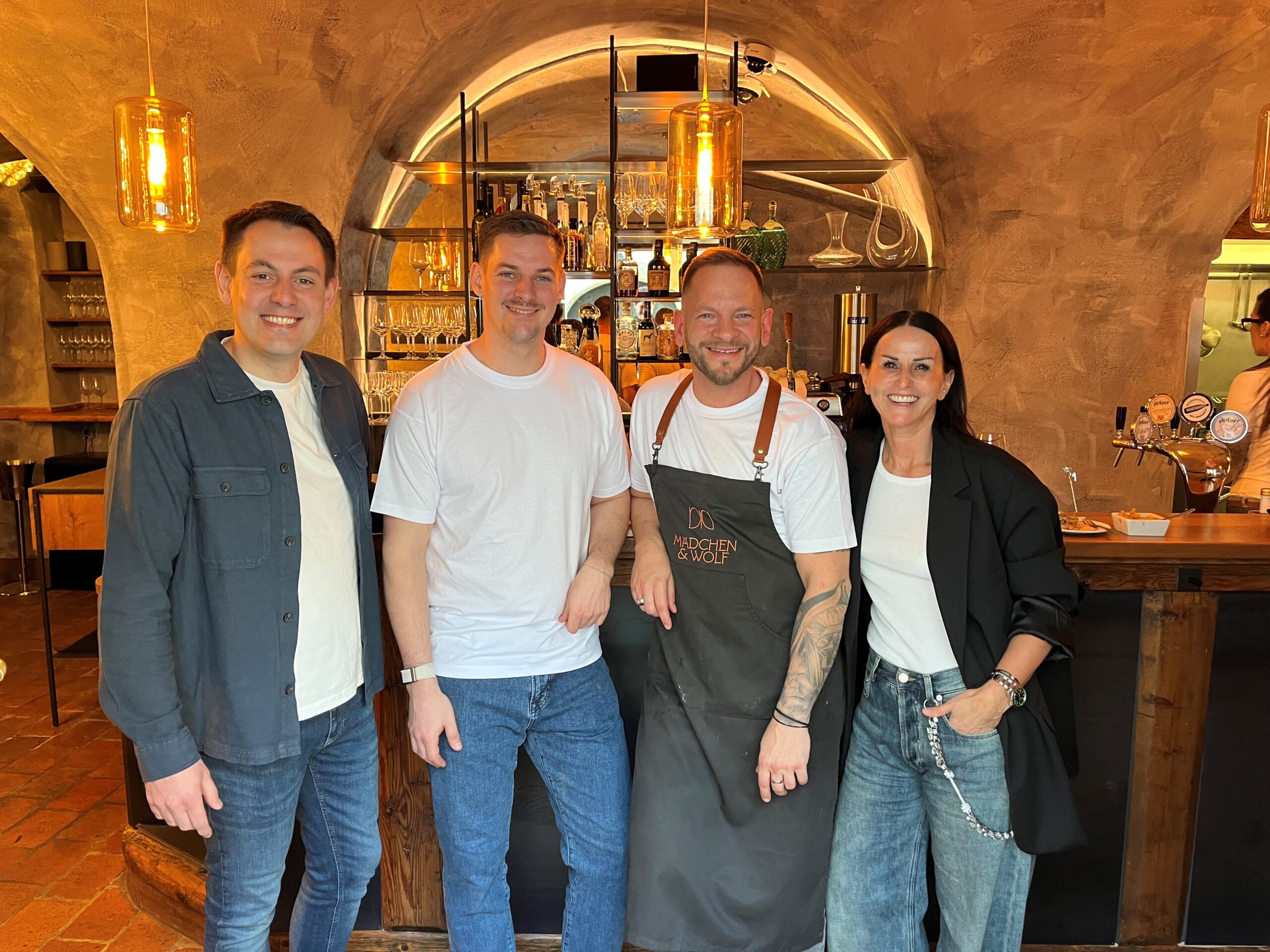 Vier lächelnde Personen stehen vor einer stilvoll beleuchteten Bar in einem Lokal mit Gewölbedecke. Ein Mann in der Mitte trägt eine dunkle Schürze mit dem Logo ‚Mädchen & Wolf‘. Die Atmosphäre wirkt modern und einladend.