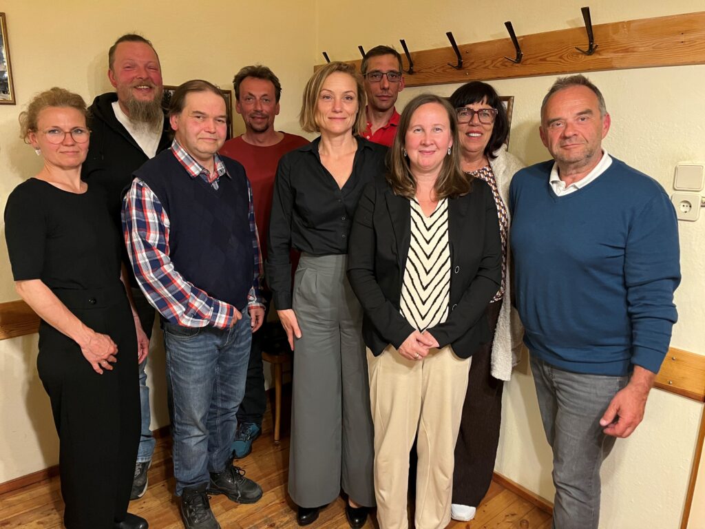 Gruppenfoto von neun Personen in einem Raum mit hellen Wänden und einer hölzernen Garderobenleiste im Hintergrund. Die Teilnehmer sind in herbstlicher Alltagskleidung (Pullover, Hemden, Sakkos) gekleidet und lächeln freundlich in die Kamera.