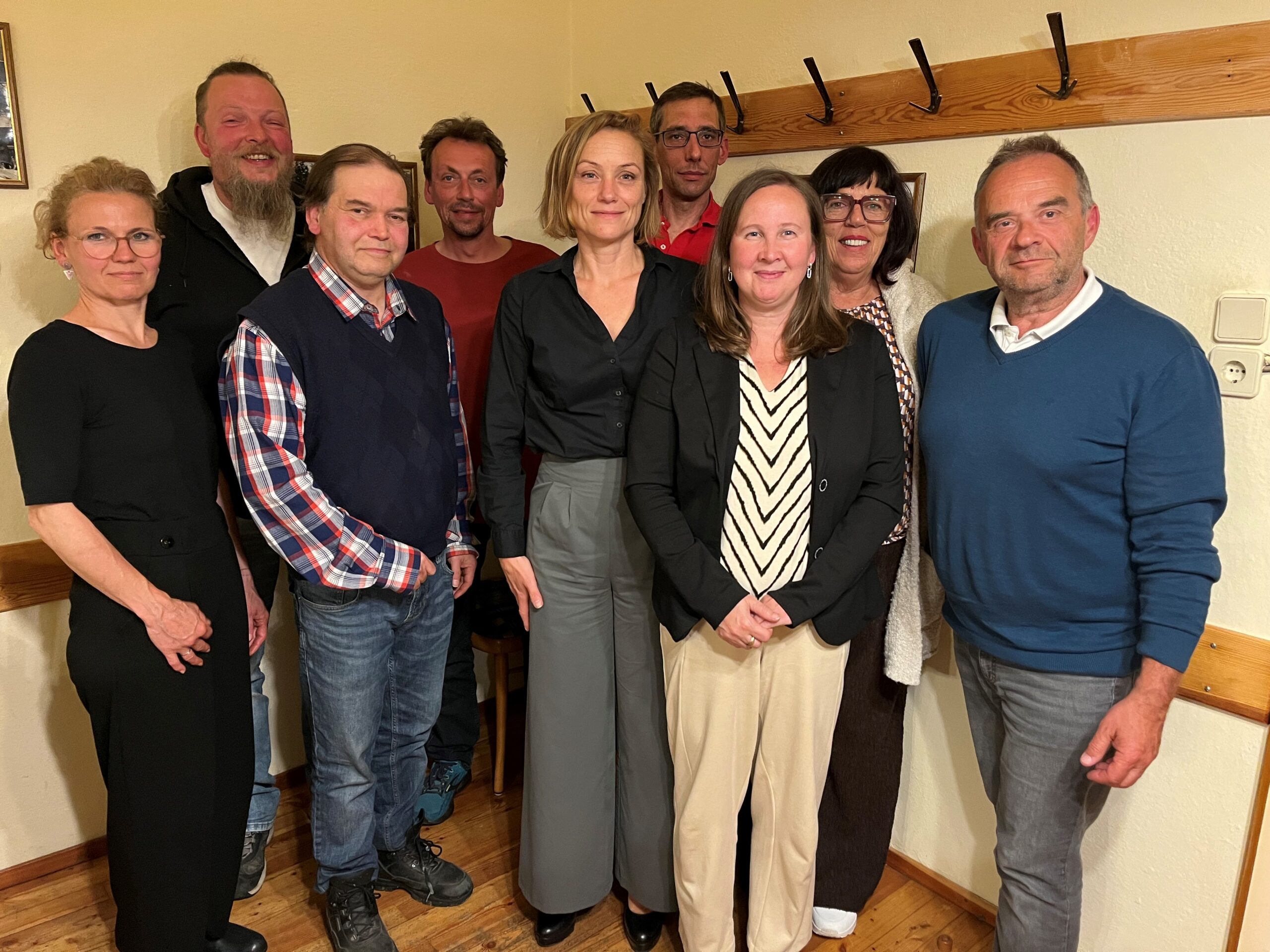 Gruppenfoto von neun Personen in einem Raum mit hellen Wänden und einer hölzernen Garderobenleiste im Hintergrund. Die Teilnehmer sind in herbstlicher Alltagskleidung (Pullover, Hemden, Sakkos) gekleidet und lächeln freundlich in die Kamera.