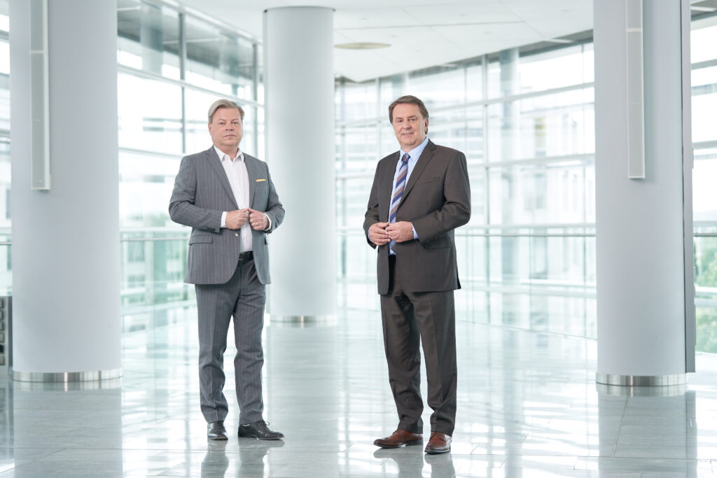 Harald Servus im grauen Anzug links vor einer Glasfront, rechts neben ihm im dunklem Anzug Wolfgang Ecker