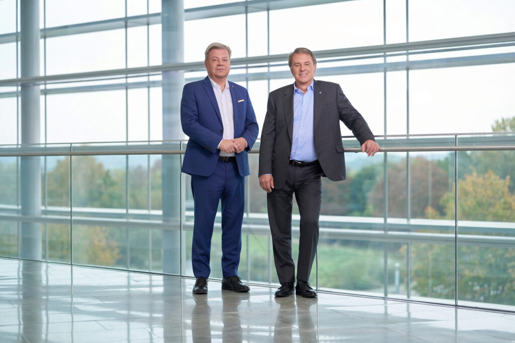 Harald Servus links im blauem Anzug und Wolfgang Ecker im dunklem Anzug vor einer Glasfront im Hintergrund Natur