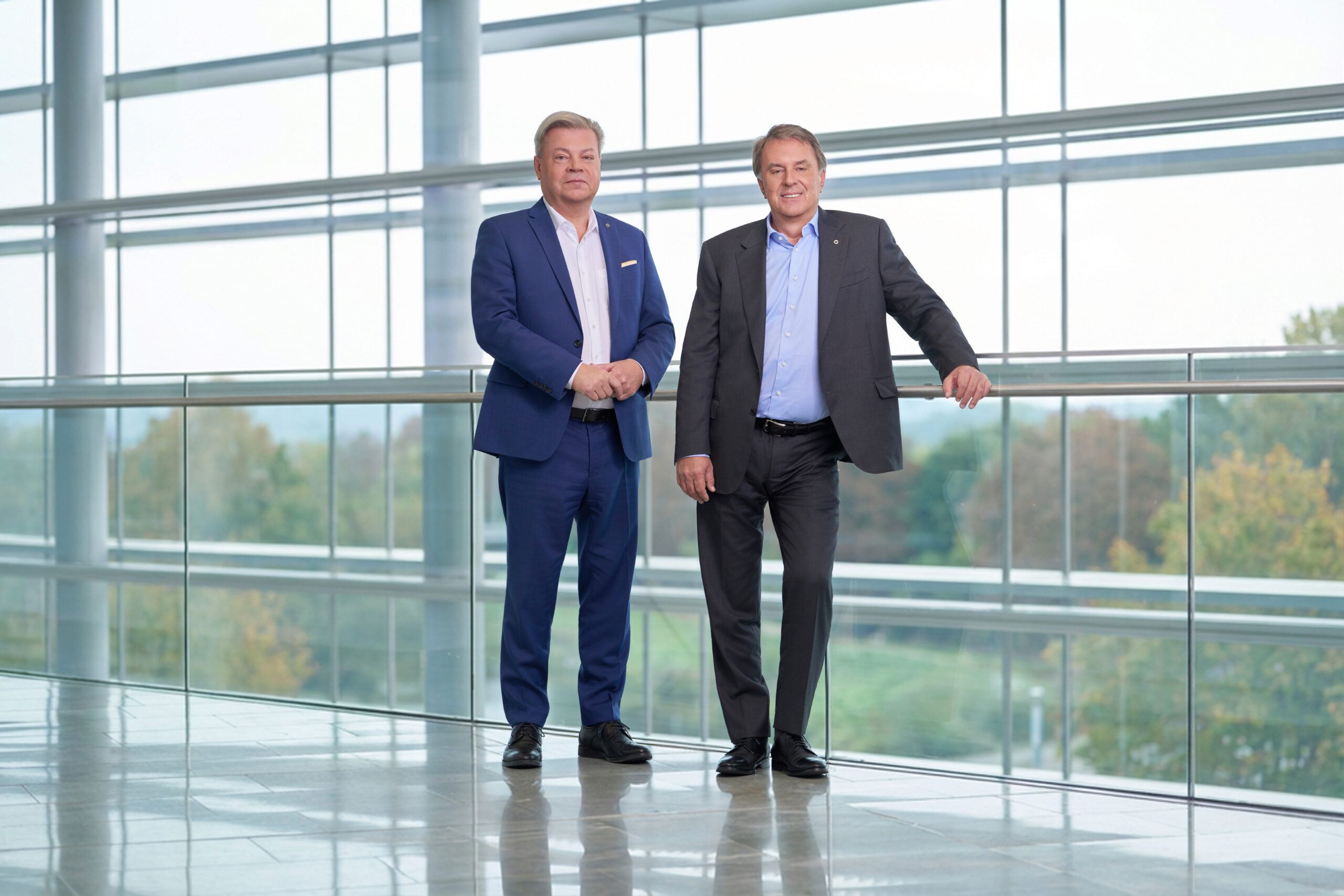 Harald Servus links im blauem Anzug und Wolfgang Ecker im dunklem Anzug vor einer Glasfront im Hintergrund Natur