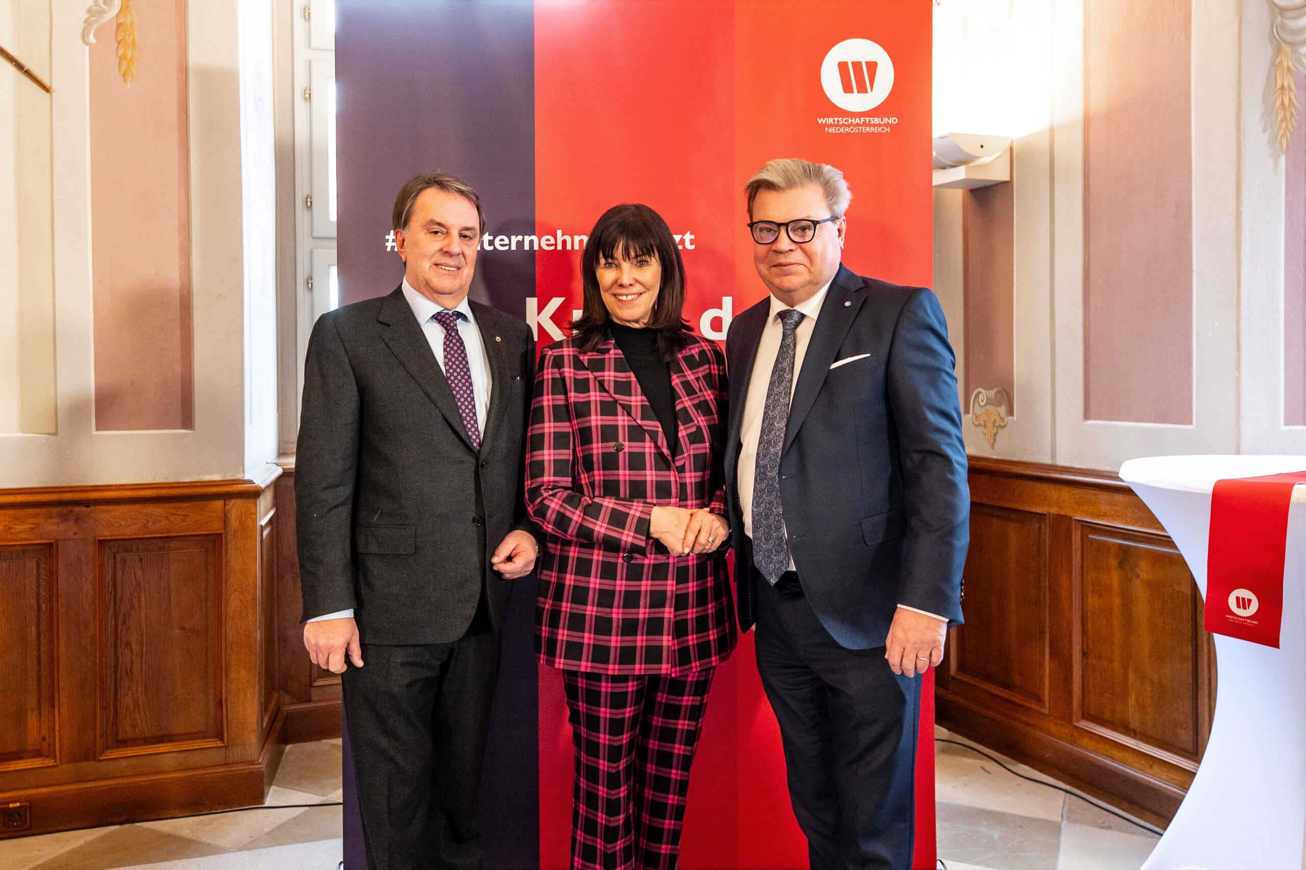 Auf dem Bild Wolfang Ecker in dunklem Anzug, Martha Schulz in der Mitte mit Muster Blazer und Hose sowie Harald Servus links vor einem Roll Up des Wirtschaftsbundes Niederösterreich