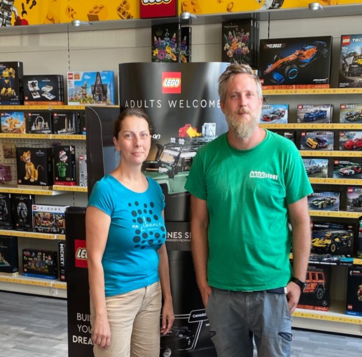 Zwei Personen, ein Mann im grünen "Brick Store"-T-Shirt und eine Frau in einem blauen Oberteil, stehen lächelnd in einem Spielwarengeschäft vor einem Regal voller LEGO-Sets und einem großen Werbeaufsteller.