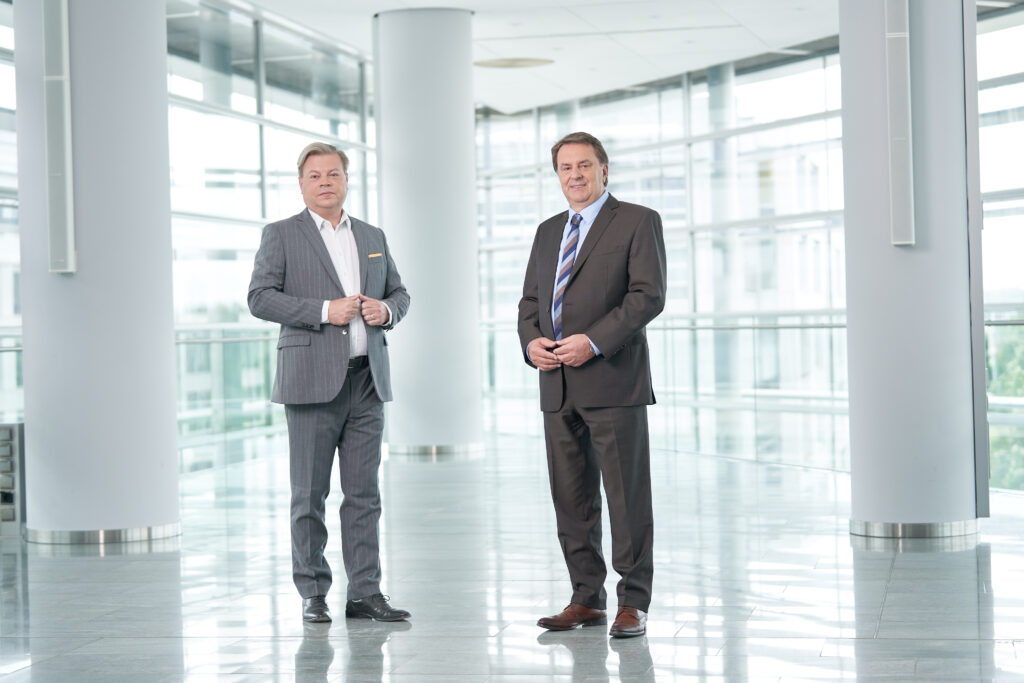 Harald Servus im grauen Anzug links vor einer Glasfront, rechts neben ihm im dunklem Anzug Wolfgang Ecker